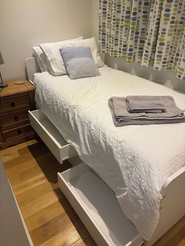 A neatly made bed with a white duvet, a grey towel, and a grey pillow. There is a wooden nightstand with a lamp and a drawer. The room has wooden flooring and a white wall. - St Matthias Lodge - Delightful 2 bed cottage (Torquay)