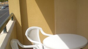 Un petit balcon avec deux chaises en plastique blanches et une table blanche, d’où l’on peut voir une rue et des voitures garées.