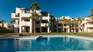 A swimming pool with a clear blue water surface, surrounded by a building with multiple balconies and palm trees. - Lovely 2 bedroom,2 bathroom ground floor apartment adjacent to pool Sleeps 4/5. (San Javier)