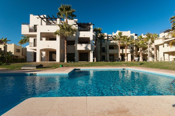 A swimming pool with a clear blue water surface, surrounded by a building with multiple balconies and palm trees.