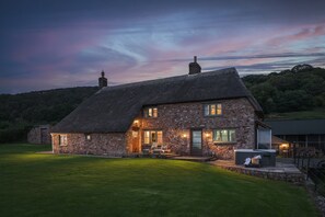 Une maison en pierre avec un toit de chaume, une entrée bien éclairée et un patio aménagé avec des sièges d’extérieur.