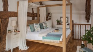 Uma cama com dossel e cortina, vigas de madeira, uma mesinha de cabeceira branca, uma mesa de apoio com abajur, uma pequena planta e uma estante de livros.