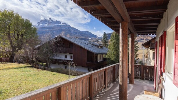 Un espacio al aire libre techado con una terraza de madera, una mesa con una silla y vistas a montañas y edificios al fondo.