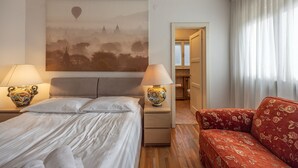 Habitación de hotel con cama, dos lámparas de noche, un sofá, un cuadro de un paisaje y un baño visible a través de una puerta abierta.