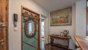 Un pasillo con una puerta de madera, un espejo, un cuadro y una mesa con un florero y un objeto enmarcado.