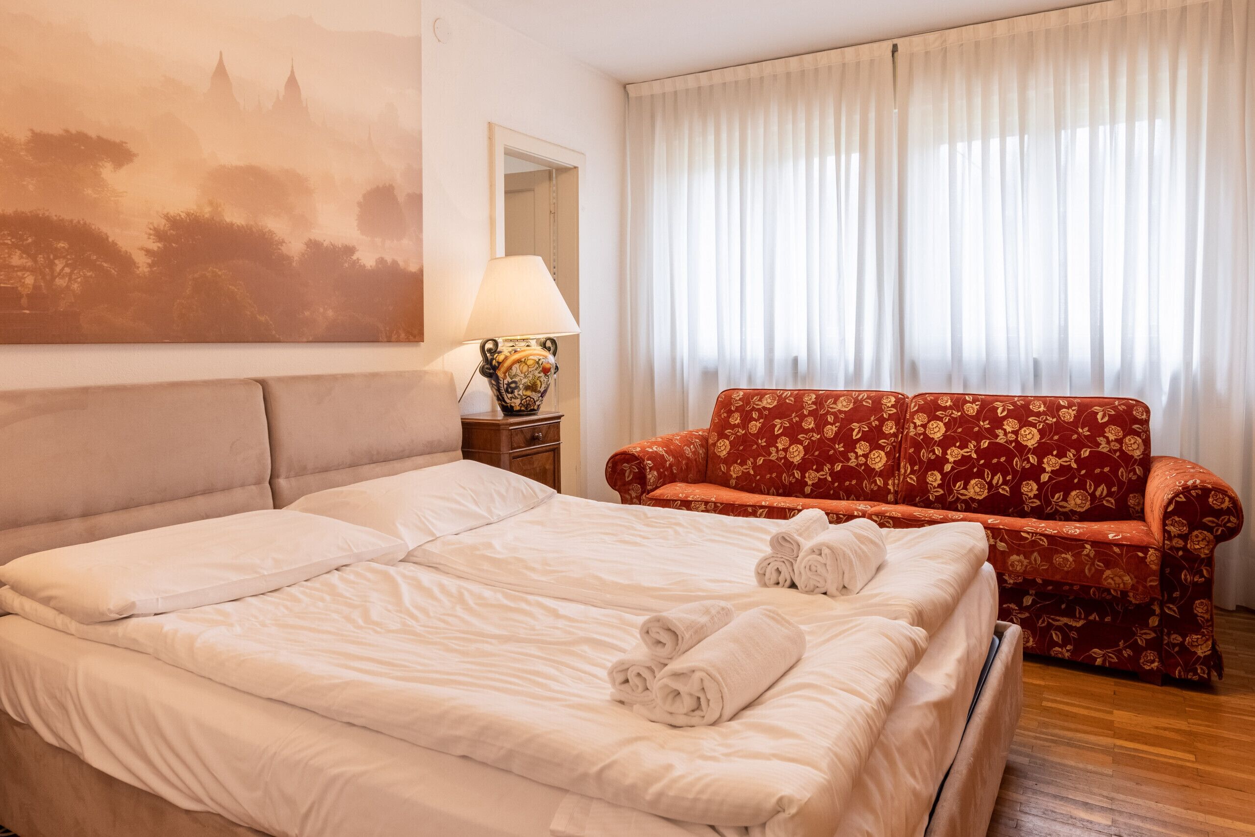 Una habitació d'hotel amb un llit, un sofà floral vermell, una làmpada i un quadre a la paret.