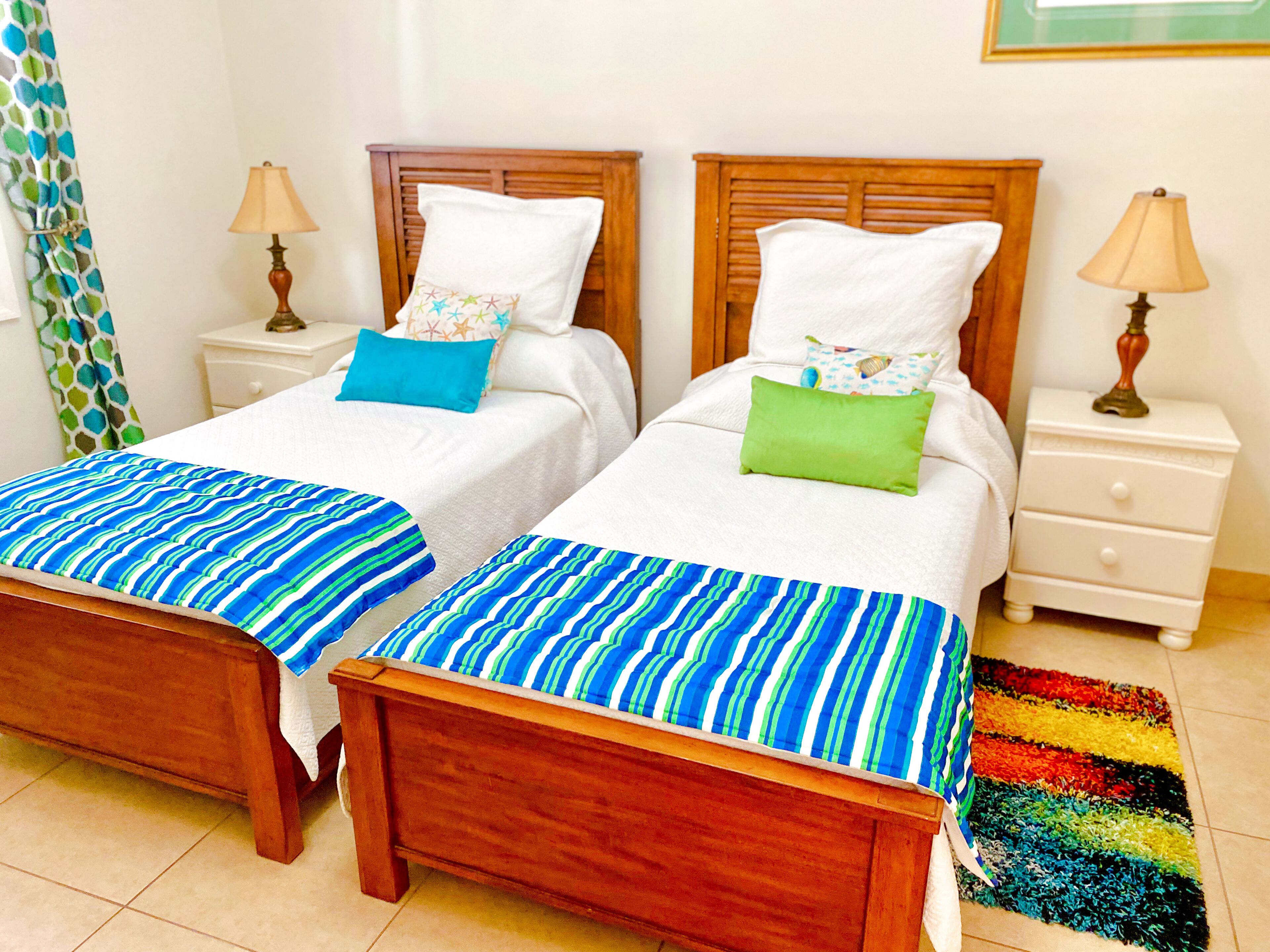 A double room with two wooden beds, each with a white duvet and striped bedspreads. There are two bedside tables with lamps, a colorful rug, and a curtain with a leaf pattern.