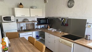 Uma cozinha compacta com micro-ondas, torradeira, chaleira e fogão. Há uma mesa de jantar com cadeiras, um relógio na parede e armários.
