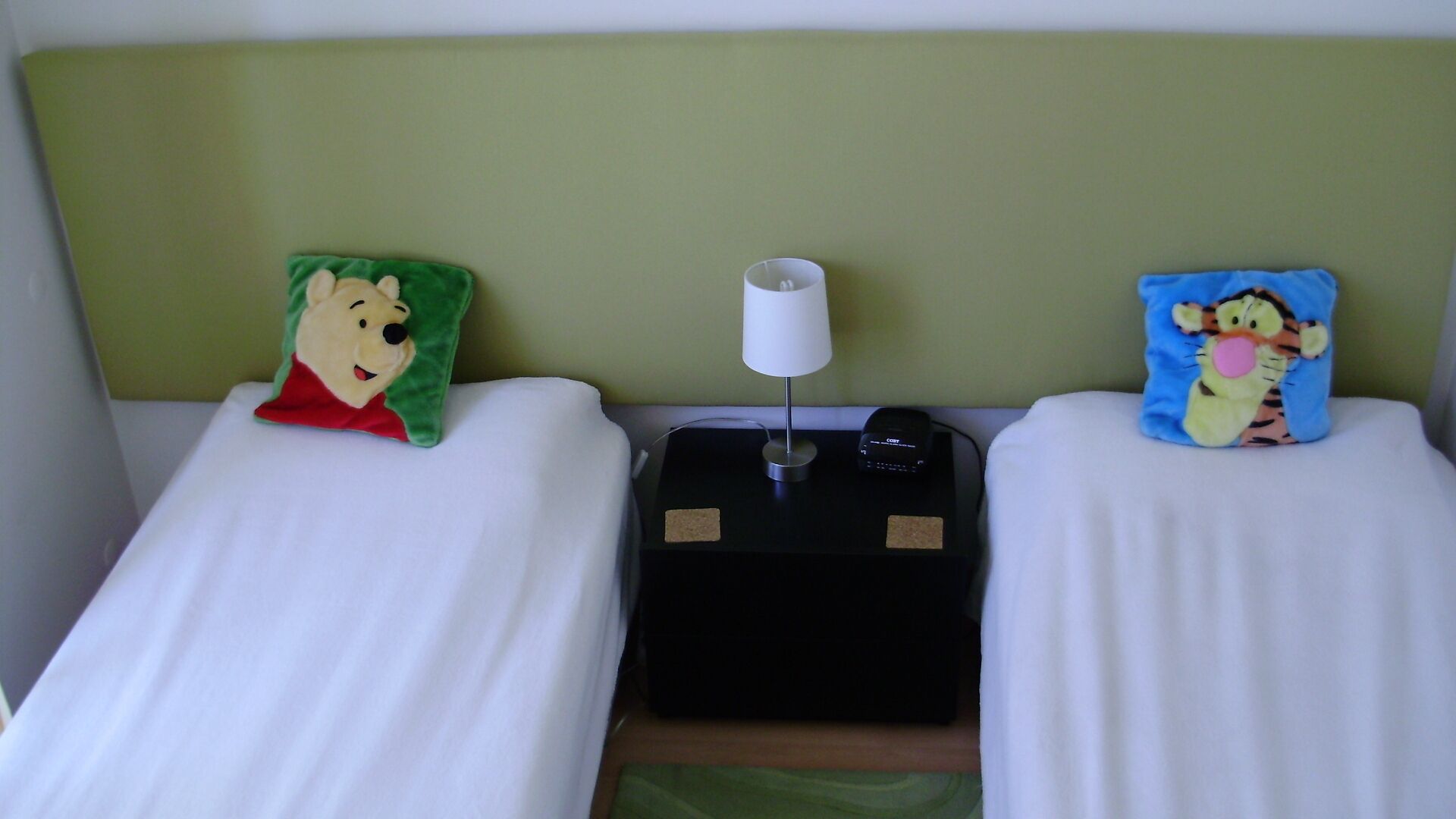 Two beds with green and blue pillows, a bedside table with a lamp, and a black electronic device.
