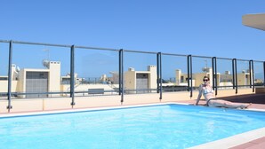 Ett poolområde med en klarblå himmel, en person som sitter vid poolen och byggnader i bakgrunden.