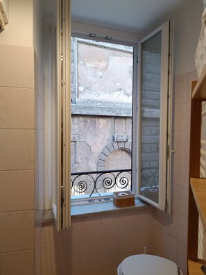 A bathroom with a toilet, a window with an open shutter, and a view of an outdoor area with a stone wall and a metal railing. - Rome: House in the heart of the 'Roman Empire (Roma)
