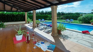 A covered outdoor area with a pool, lounge chairs, and a wooden table with a plant, wine glasses, and a book. - VILLA MARIA - RUSTIC STAY (Pollensa)