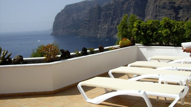 A terrace with white lounge chairs, a tiled floor, and a view of the ocean and cliffs. - Prime Location, Htd Pool, Breathtaking Sea Views nr Beach/Marina, A/C WiFi UKTV (Los Gigantes)