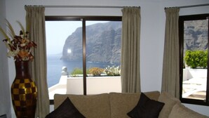 A living room with a beige sofa, two dark cushions, a checkered vase, and a view of the sea and cliffs through large windows. - Prime Location, Htd Pool, Breathtaking Sea Views nr Beach/Marina, A/C WiFi UKTV (Los Gigantes)
