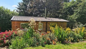 Une cabane en bois avec un toit en tuiles, entourée d’un jardin où poussent différentes fleurs et plantes.