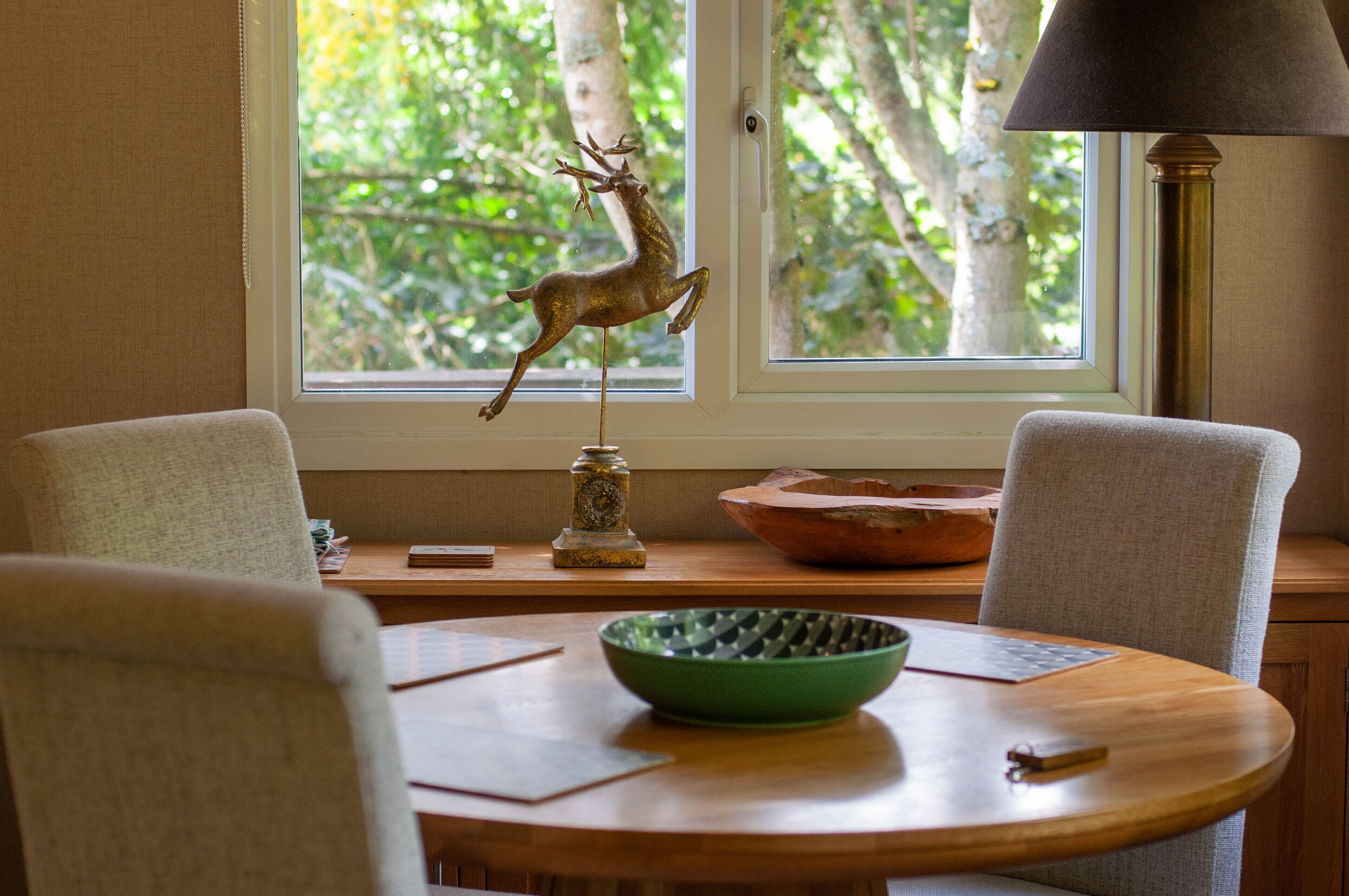 Uma sala de jantar com uma mesa redonda, cadeiras, uma tigela verde, uma escultura de veado e uma janela com vista para árvores.