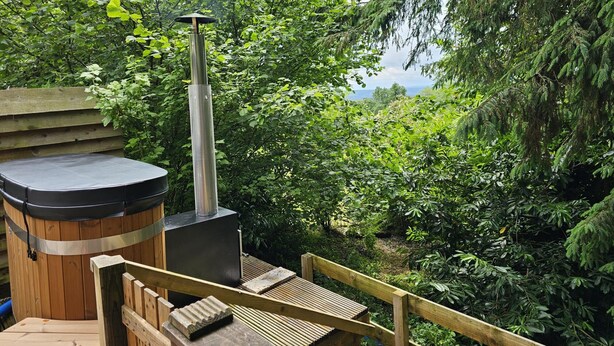 Un bain à remous doté d’une terrasse en bois et d’une cheminée en métal, entouré d’une végétation luxuriante et offrant une vue sur le paysage.
