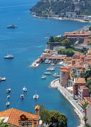 A coastal town with a marina, buildings with red-tiled roofs, and a clear blue sea.