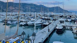 Uma marina com vários barcos atracados, um cais com um lampião e um cenário montanhoso ao fundo.
