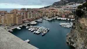 Uma marina com barcos atracados, rodeada por edifícios e uma paisagem montanhosa ao fundo.