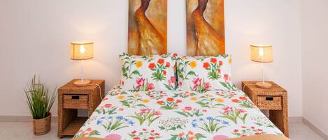 A bedroom with a floral bedspread, two wall-mounted paintings, and two bedside lamps on wooden nightstands.