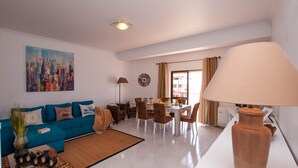 A living room with a blue sofa, a dining table with chairs, a large vase lamp, and a cityscape painting on the wall. - Walk to Beach & Strip Stylish 2BR w/ Pool, Terrace & BBQ | Albufeira (Albufeira)
