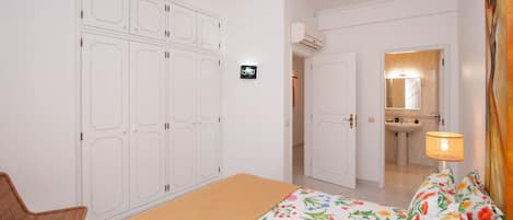 Un dormitorio con cama, una silla, un armario y un baño visible a través de una puerta abierta.