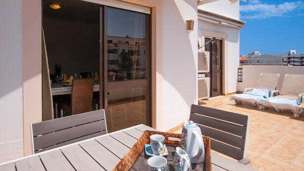 A sunlit outdoor seating area with a table set for two, a sliding glass door leading to a room, and a poolside lounge with sun loungers. - Walk to Beach & Strip Stylish 2BR w/ Pool, Terrace & BBQ | Albufeira (Albufeira)