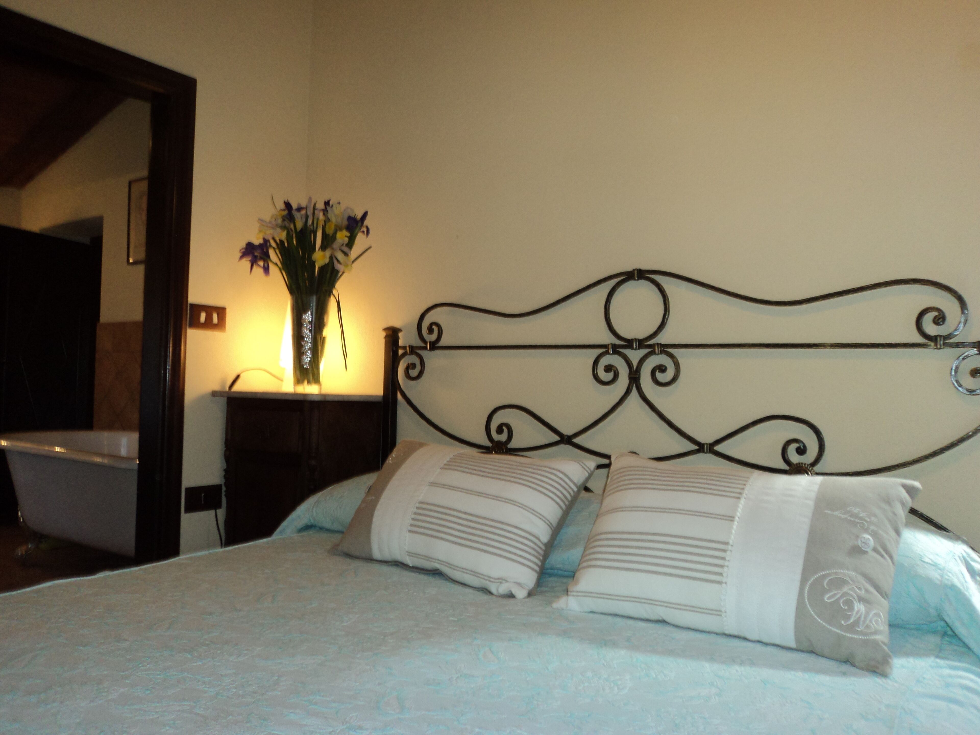 Una cama con cabecera metálica, dos almohadas y un jarrón con flores sobre una mesita de noche.