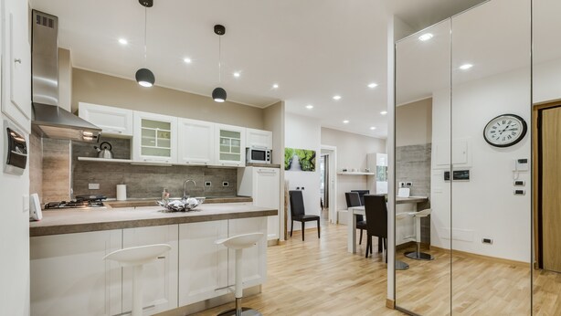 A modern kitchen with white cabinets, a stainless steel range hood, and a wooden countertop. There are two white bar stools and a dining area with a table and chairs. - VATICAN FLAT IN ROME (Roma)