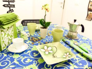 Ein Tisch gedeckt mit einer grün-weißen Tischdecke mit Blumenmuster, einer weißen Kaffeekanne und einem grünen Blumenvasen mit Pflanze.