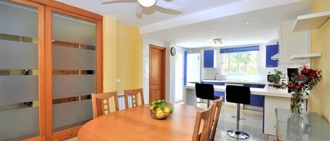 A dining area with a round wooden table, chairs, a ceiling fan, and a kitchen in the background.