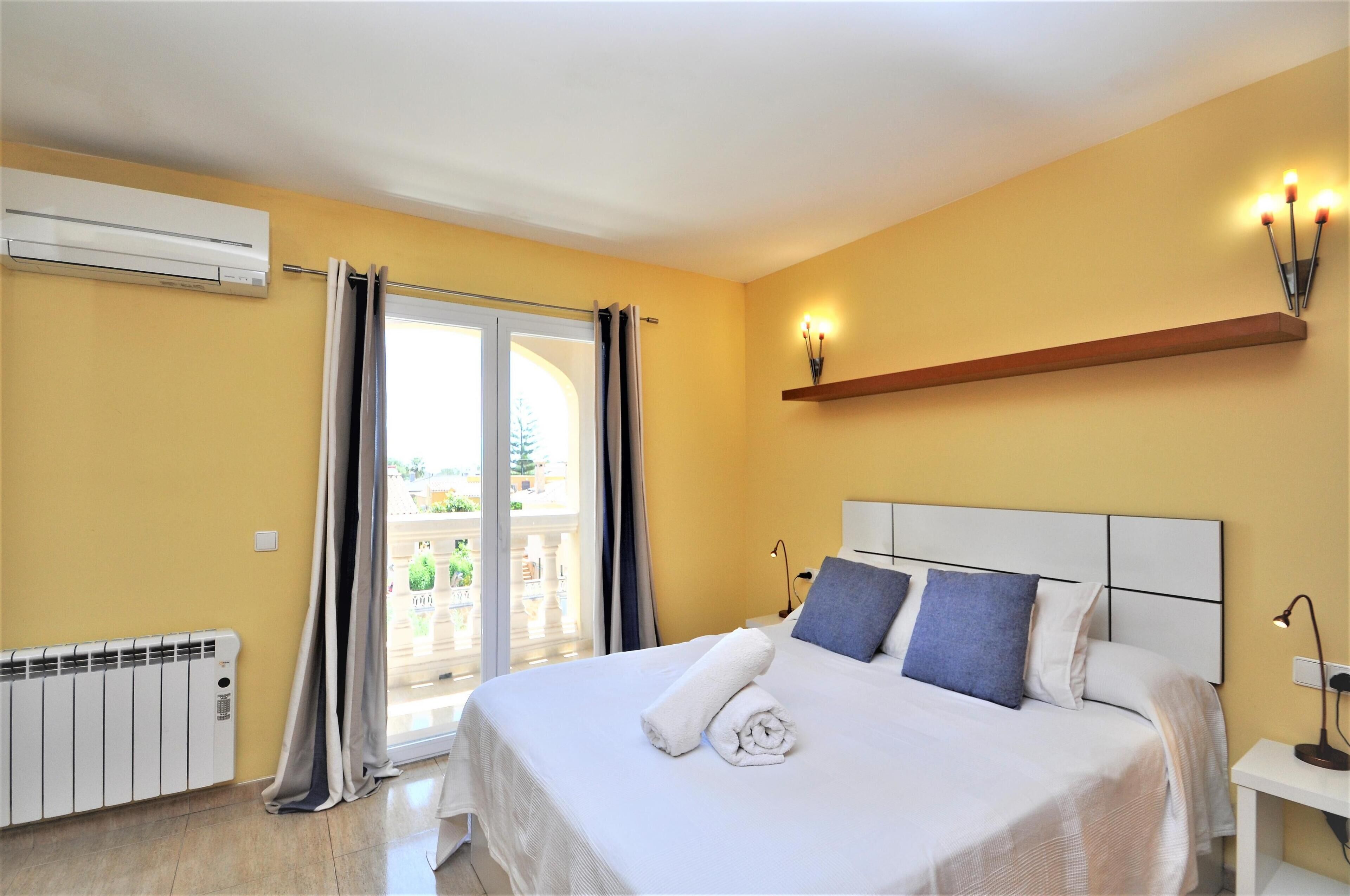 A hotel room with a bed, a window with curtains, an air conditioning unit, a radiator, and a nightstand with a lamp.