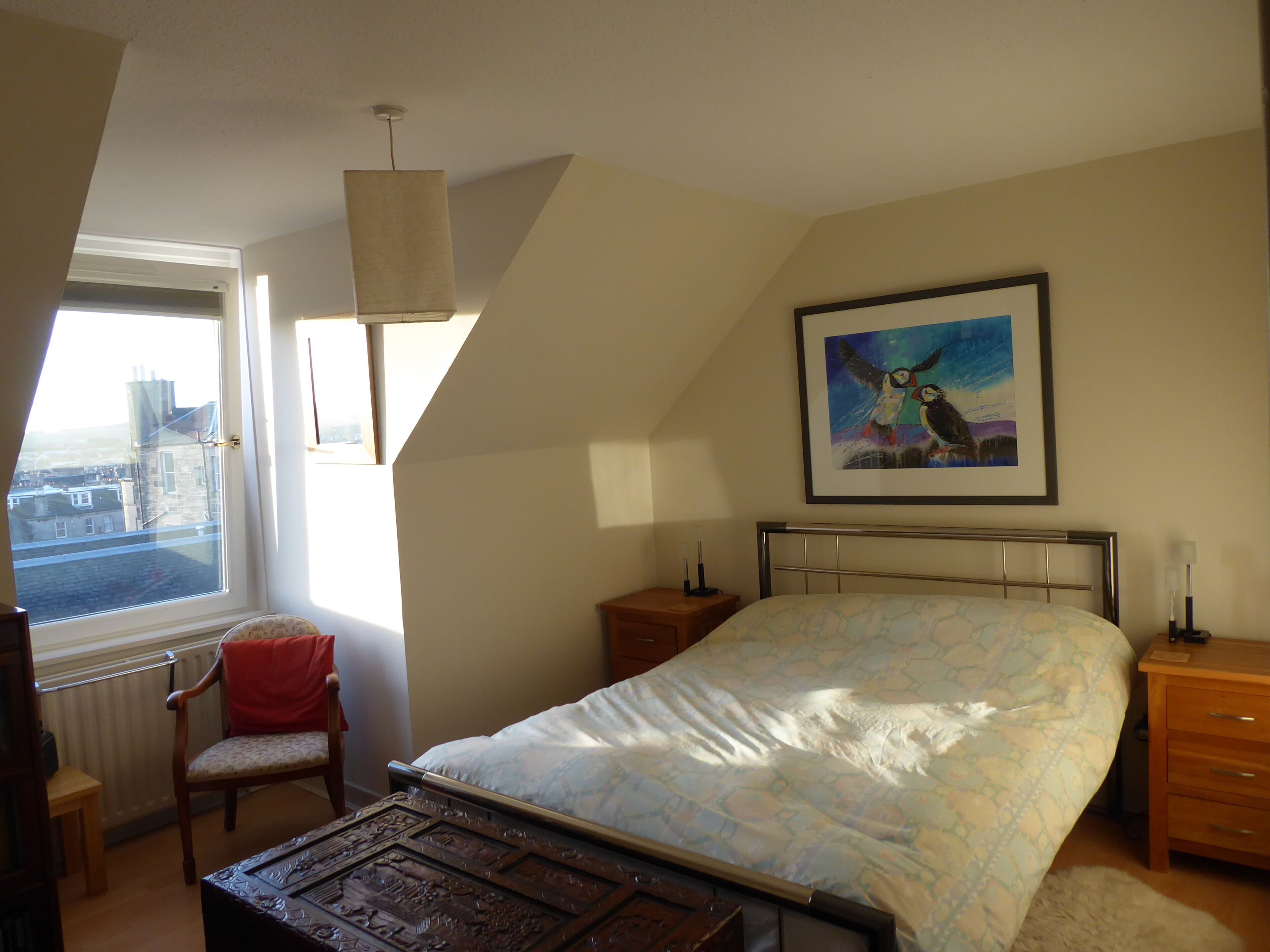A bedroom with a bed, a chair, a nightstand, a bedside table, a framed picture on the wall, and a window with a view of buildings.