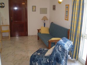 A living room with a wooden door, a sofa, a small table, and a chair. - Apartment description (Tavira)