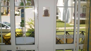 Uma porta branca com uma pequena placa, uma varanda com mesa e cadeiras, e vista para uma rua com carros estacionados e vegetação.