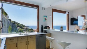 A modern kitchen with a large window overlooking a coastal view, a dishwasher, and a bar area with a TV. - Luxury villa apartment with panoramic sea views and small private garden (Nice)