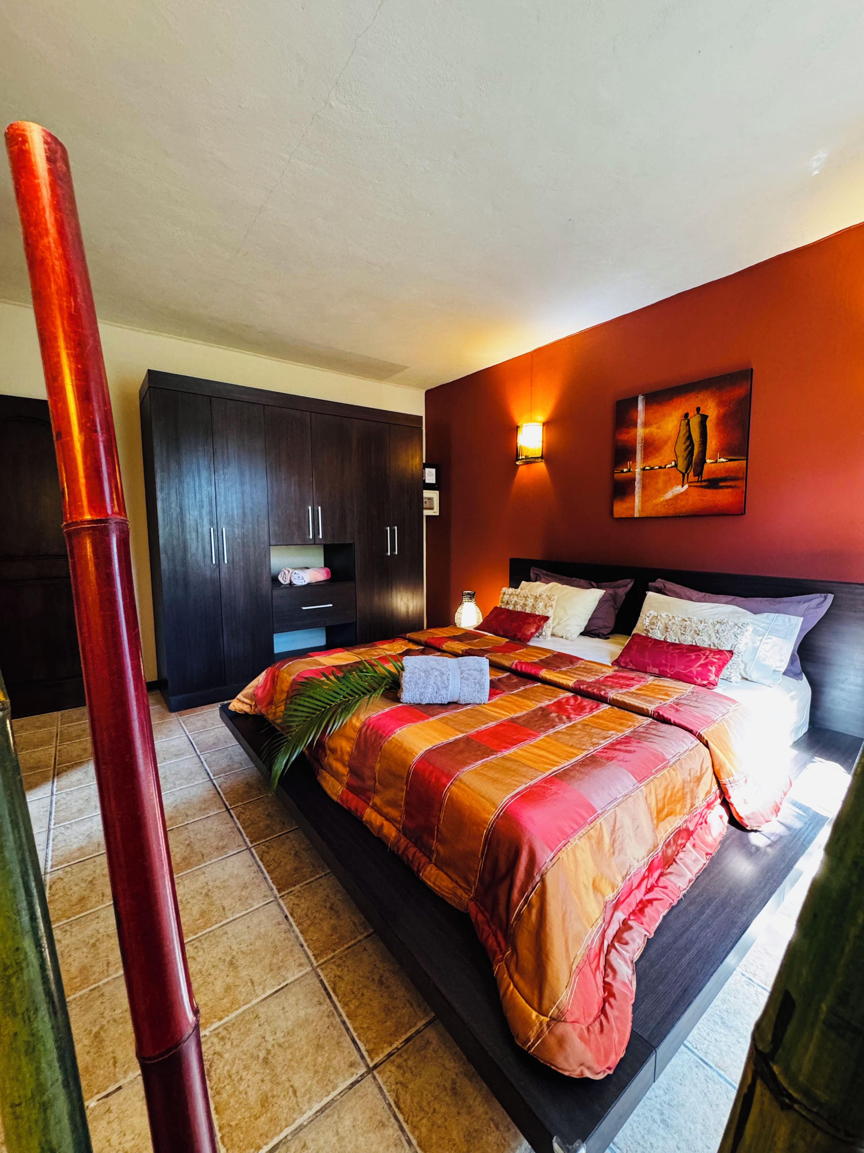 A bedroom with a bed, dark wooden wardrobe, a painting on the wall, and a red pillar.