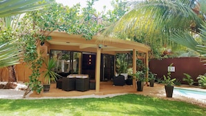 Un área de estar al aire libre cubierta con piscina, rodeada de abundante vegetación y palmeras.