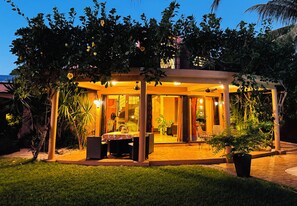 A covered outdoor seating area with tables and chairs, surrounded by lush greenery and a view of the sea.