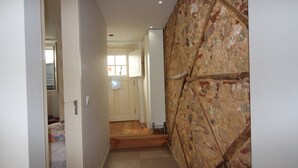 Un pasillo estrecho con piso de madera, pared de piedra a la derecha y una puerta que da a una habitación con cama.