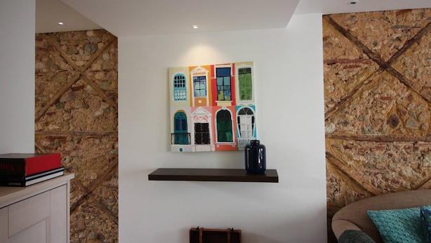 Una habitación con pared de piedra, una obra de arte colorida, una estantería con un libro y un jarrón, y un sofá con un cojín estampado.