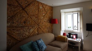 Una sala de estar con una pared de piedra, un sofá con cojines estampados, una lámpara roja, un escritorio con una computadora y una ventana con vistas.