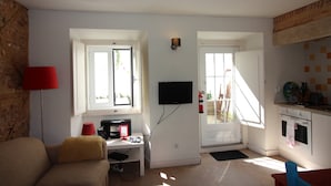 Un espacio habitable compacto con sofá, una mesita, televisor montado en la pared, zona de cocina con estufa y armarios, y una puerta que da al exterior.