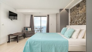 Una habitació d'hotel moderna amb un llit gran, un televisor fixat a la paret, una taula petita i vista al mar per la finestra.