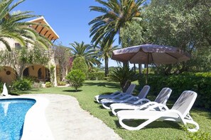 Un espace aménagé au bord de la piscine, avec des chaises longues et un grand parasol, entouré de palmiers et d’un bâtiment doté d’arcades.