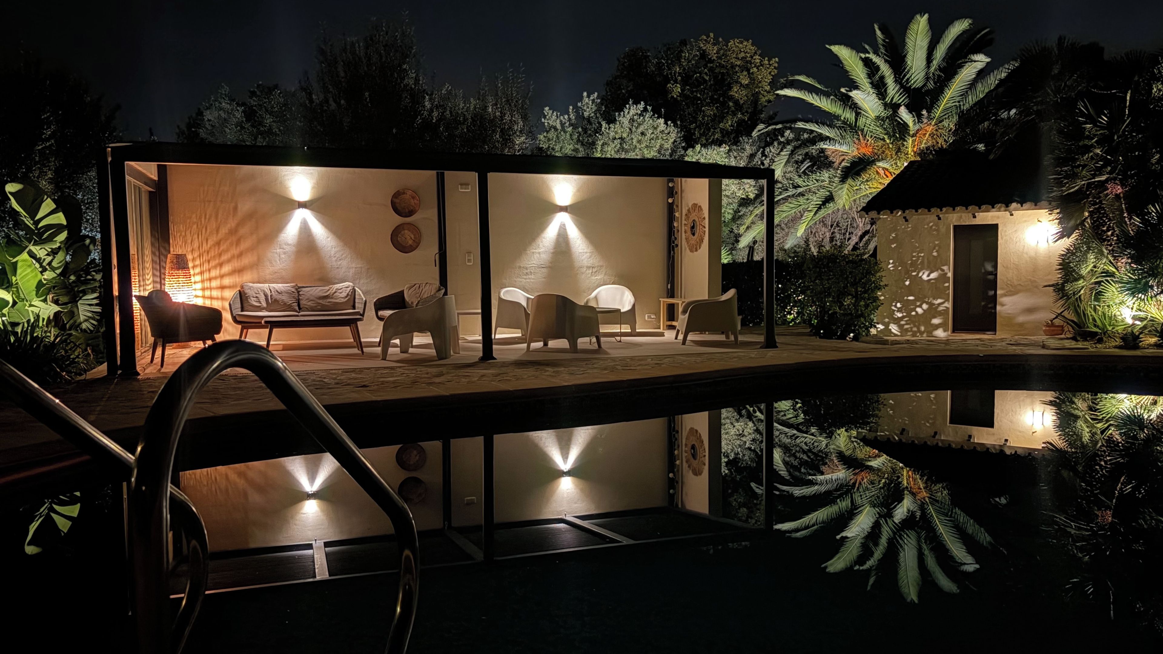 An outdoor seating area with a pool, surrounded by palm trees and a well-lit interior.