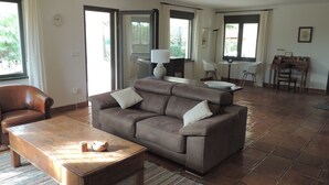 Een ruime woonkamer met een bruine leren bank, een houten salontafel en een eethoek met een kleine tafel en stoelen.