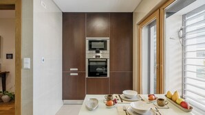A modern kitchen with a built-in oven and microwave, a dining table set with plates, bowls, and fruits, and a window with blinds. - Sunny Central Luxury, 2 bedroom/2WC, garage, don't need car, 8 min walk beach (Cascais)