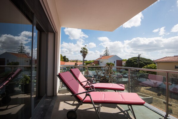 Un balcon avec deux fauteuils de jardin à rayures rouges, une rambarde en verre et une vue sur le quartier.